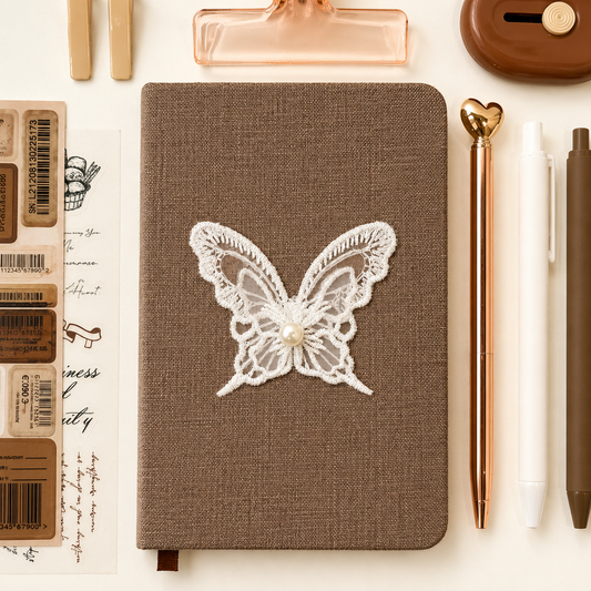 Brown notebook with a lace butterfly design on a white surface with stationery items.