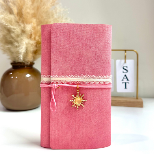 Pink notebook with decorative elements on a white surface with a vase and dried plants in the background.