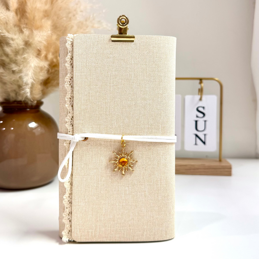 Beige notebook with a decorative clip and sun charm on a white surface with a vase and calendar in the background.