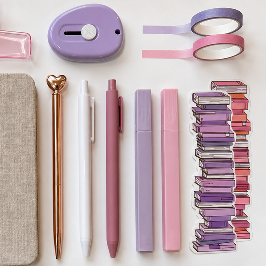 Pastel stationery essentials arranged in a minimal flat lay featuring pink and lavender washi tape, aesthetic pens and highlighters, a rose gold heart-top pen, mini label maker, linen notebook cover, and stacked book illustration sticker on a soft white background.