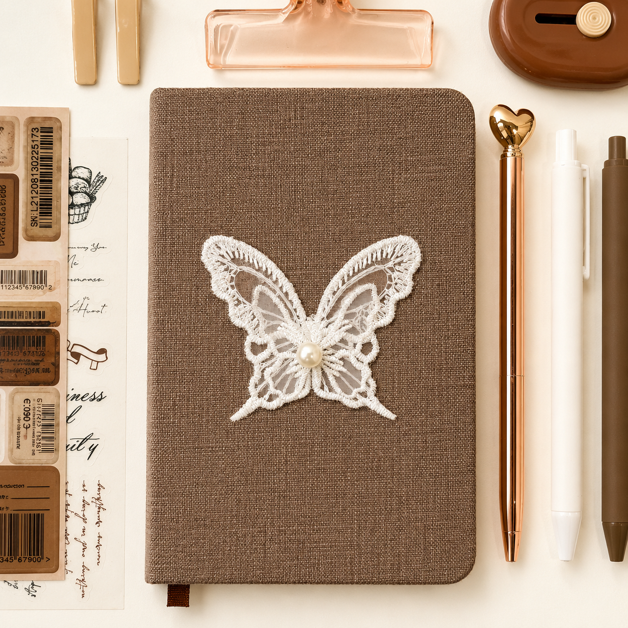 Brown notebook with a lace butterfly design on a white surface with stationery items.