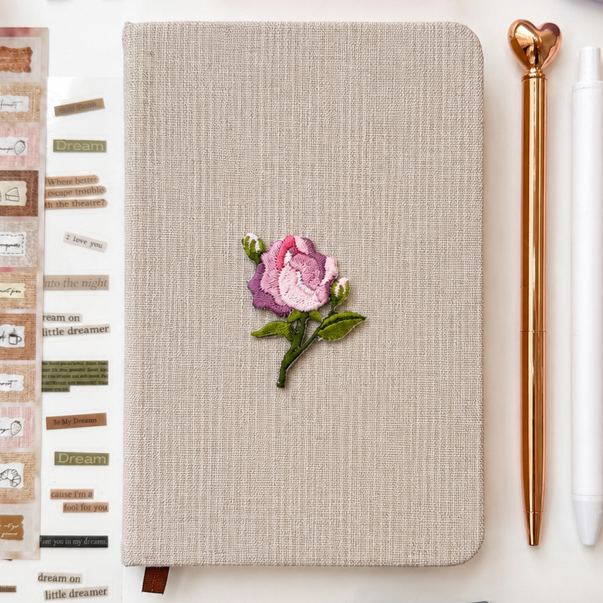 Minimal aesthetic linen journal with embroidered pink floral detail styled beside neutral scrapbook stickers and elegant rose gold and white pens on a soft white background, creating a cozy vintage-inspired stationery flat lay.