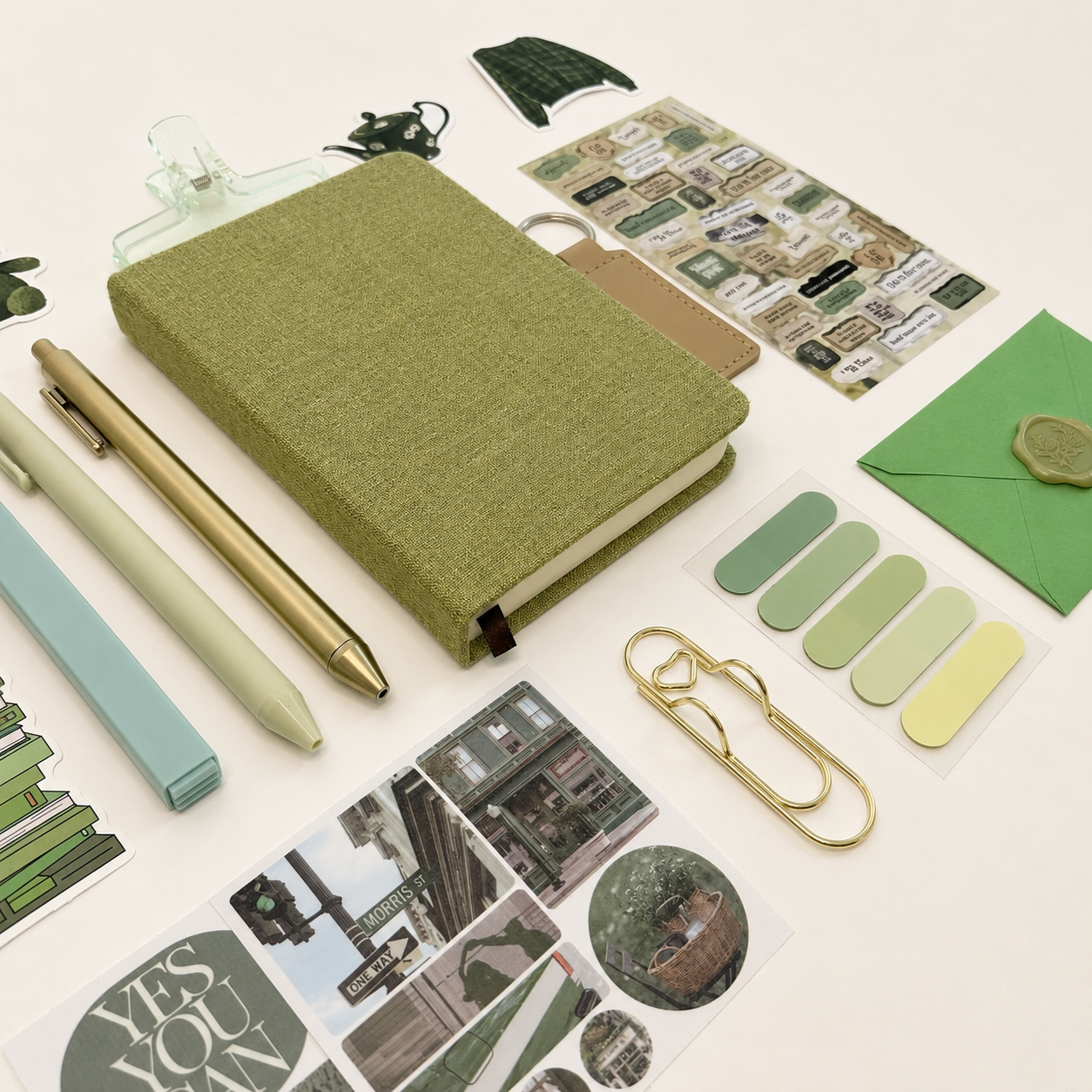 Collection of stationery items including a green notebook, pens, stickers, and paperclips on a white background.