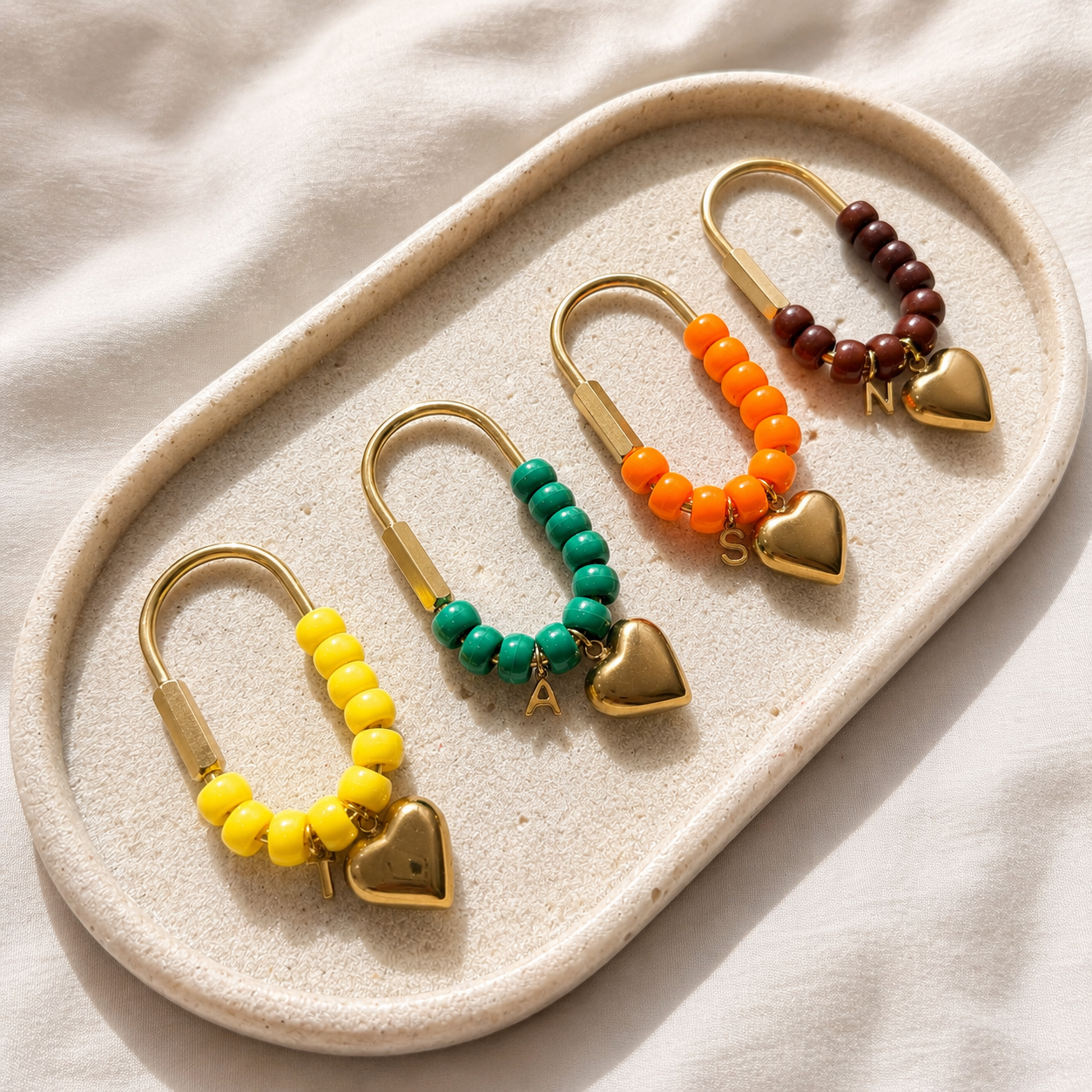 Personalized beaded heart keychains with gold hardware displayed on a textured cream tray. Each handmade keychain features colorful acrylic beads, a gold heart charm, and a custom initial charm, available in yellow, green, orange, and brown color combinations.