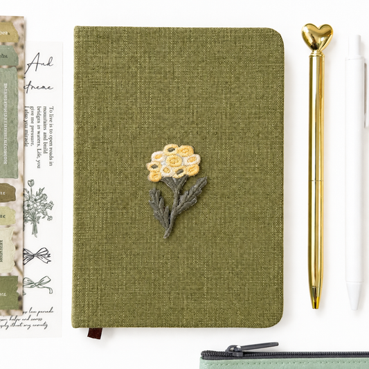 Green notebook with floral design, gold pen, and decorative item on a white background