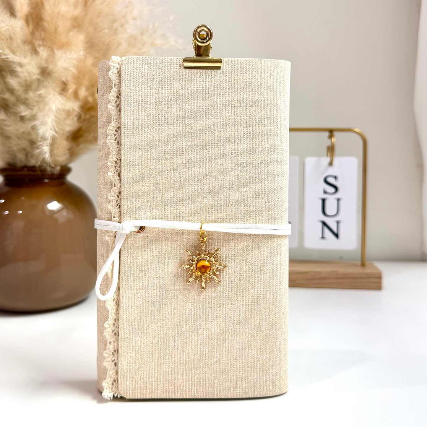 Beige notebook with a decorative clip and sun charm on a white surface with a vase and calendar in the background.