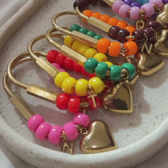Video featuring a collection of handmade personalized heart keychains in multiple vibrant colors. Each keychain is crafted with glossy beads, gold hardware, a heart charm, and a custom initial charm. The video showcases the variety of color options, fine details, and thoughtful craftsmanship against a soft neutral background