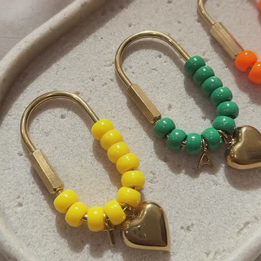 Video showcasing handmade personalized heart keychains with colorful acrylic beads and gold hardware. Each keychain features a gold heart charm and custom initial charm, displayed on a textured cream tray in yellow, green, orange, and brown color combinations. The video highlights the details, shine, and craftsmanship of each piece