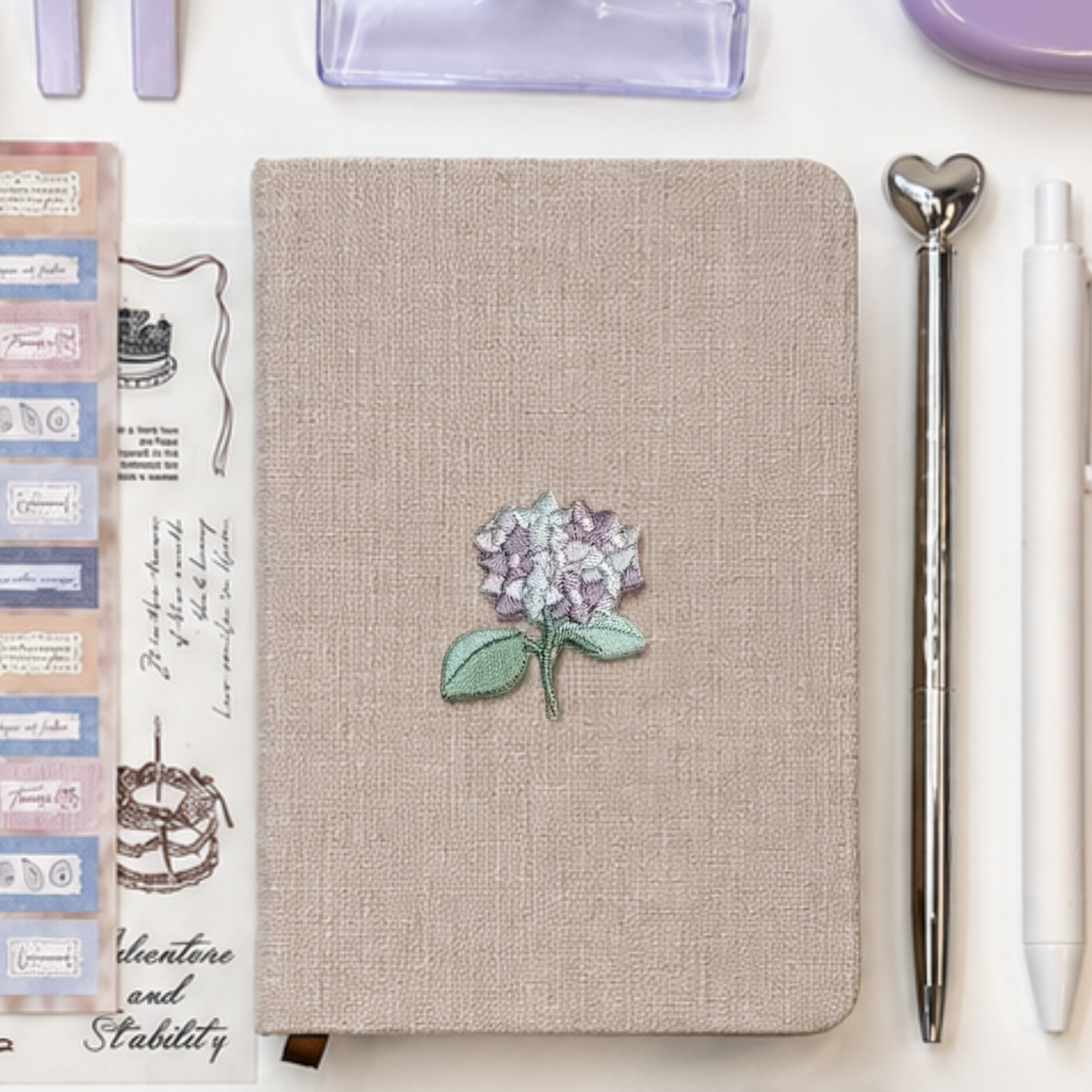 Neutral linen aesthetic journal with embroidered lavender hydrangea detail styled beside minimalist silver and white pens, pastel journaling stickers, and soft purple stationery accessories in a clean flat lay composition.
